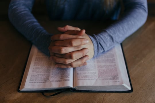Hands folded in prayer on an open Bible resting on a wooden table