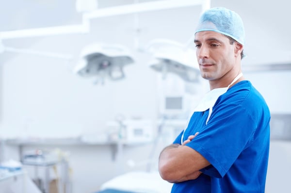 Medical professional in blue scrubs and a surgical cap standing with arms crossed in an operating room