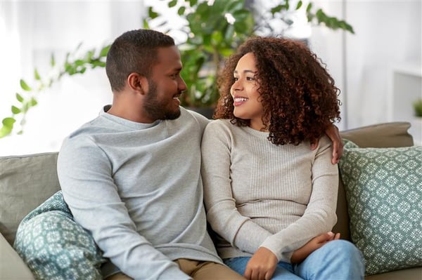 A couple sits closely together on a living‑room couch, relaxing with arms around each other as they talk in a bright, cozy home setting.