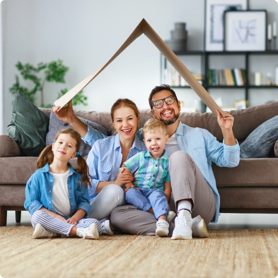 Smiling family of four sitting on a living room rug under a cardboard roof held over their heads