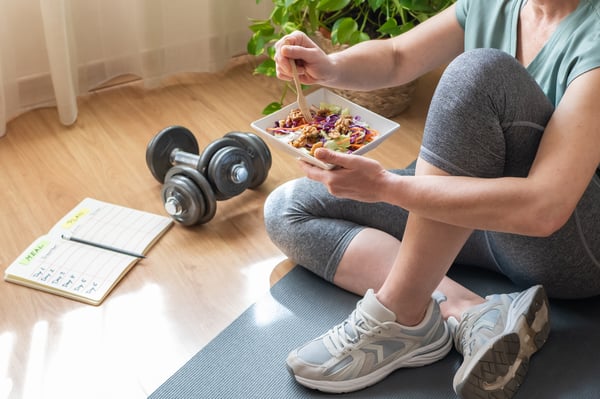 Person in workout clothes eats a salad on a yoga mat beside dumbbells and an open meal-planning notebook