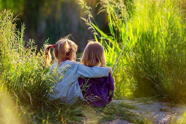 Two young girls sitting in tall sunlit grass, one with her arm around the other