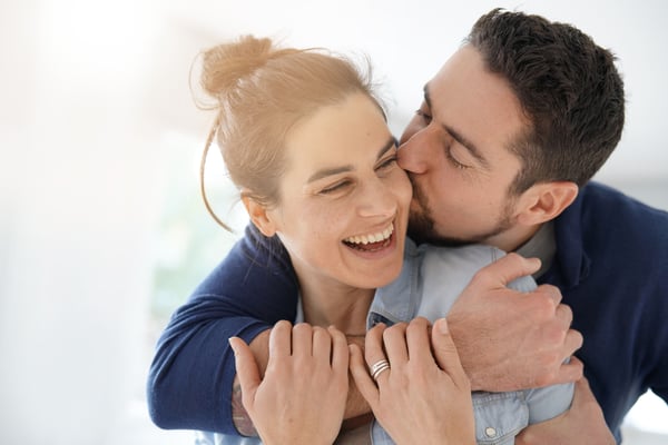 Man embraces a laughing woman from behind and kisses her on the cheek