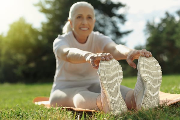 Smiling older woman stretches toward her sneakers on a yoga mat in a sunny park