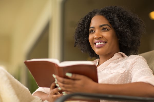 Smiling woman with curly hair relaxes in a lounge chair holding an open Bible