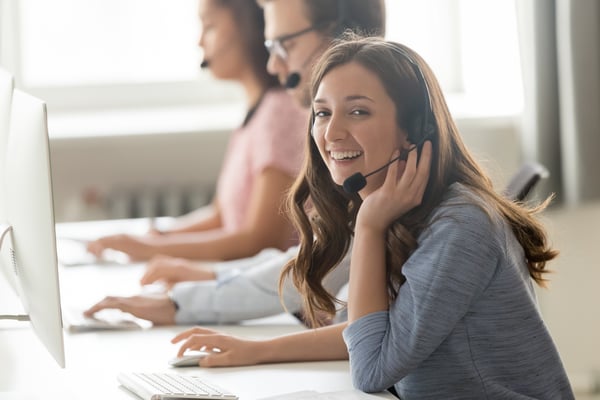 Smiling woman wearing a headset at her computer, with coworkers on headsets beside her in a call center