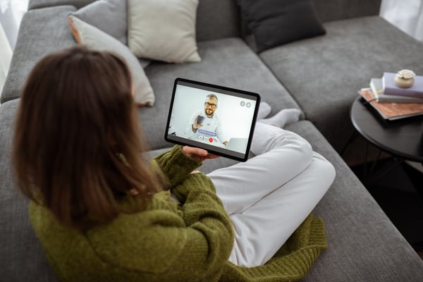 Woman on a sofa holding a tablet during a telehealth video call with a doctor
