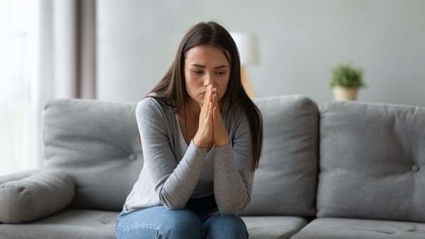 Young woman sitting on a gray couch with her hands pressed to her lips, looking worried
