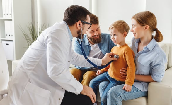 Doctor listens to a young boy's chest with a stethoscope as two smiling adults sit with him on a couch
