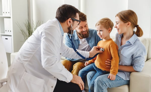 Doctor listens to a young boy's chest with a stethoscope as two smiling adults sit with him on a couch