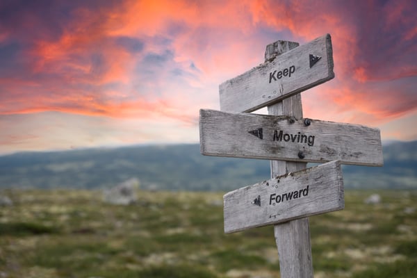 Weathered wooden signpost with arrows reading Keep Moving Forward under a pink and orange sunset sky