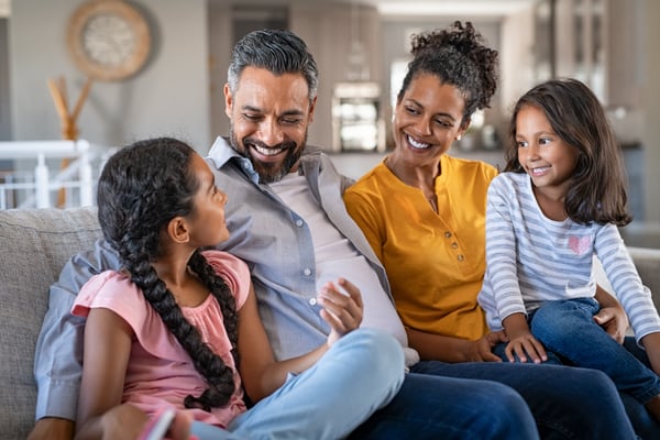 Family of four with two young girls talks and laughs together on a living room couch