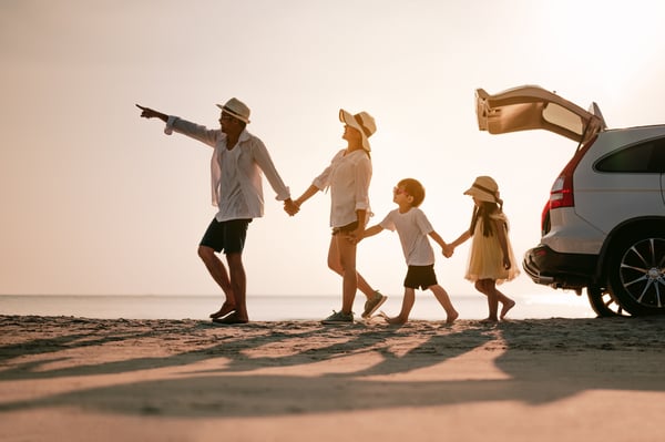 Family of four holding hands as they walk on a beach at sunset beside an SUV with its hatch open