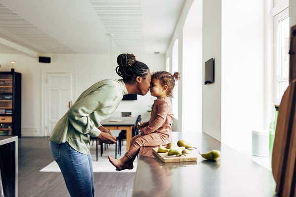 Woman touches foreheads with a toddler sitting on a kitchen counter beside fresh pears
