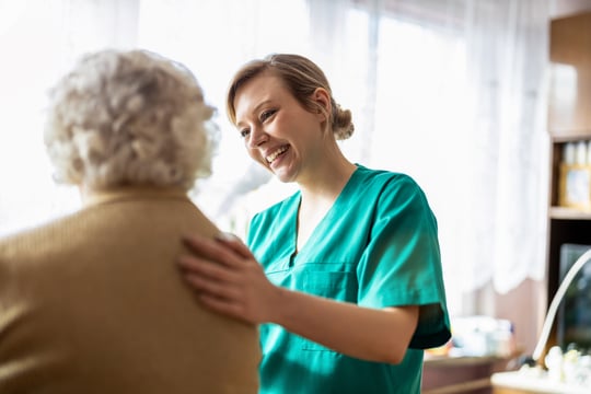 Smiling nurse in teal scrubs rests a hand on the shoulder of an older woman with curly white hair