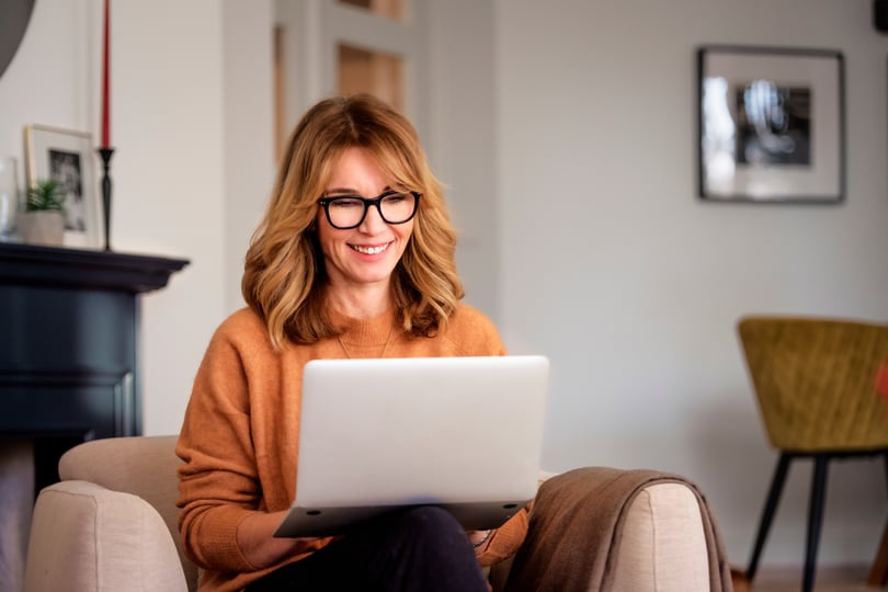 A woman wearing glasses smiling warmly as she looks at her laptop, exuding a sense of joy and focus.