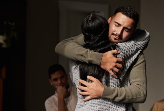 Man with eyes closed holds a woman in a comforting hug in a dim room while another man sits nearby