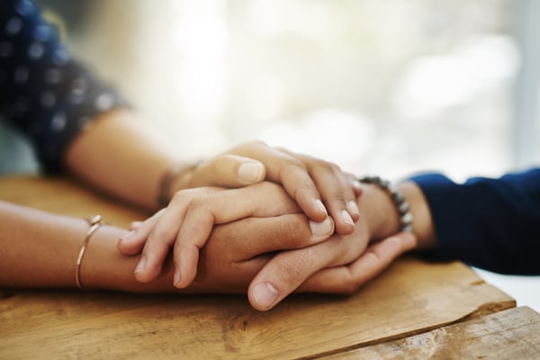 Close-up of two people holding hands on a wooden table in a gesture of comfort