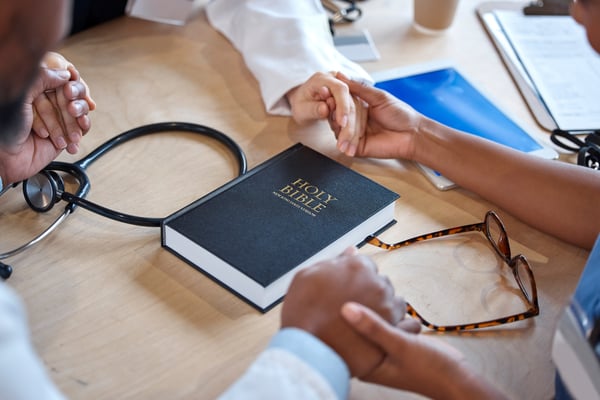 People join hands around a table with a Holy Bible, a stethoscope and eyeglasses