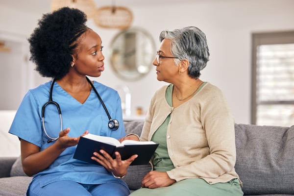 Health care worker in blue scrubs holds an open book while talking with an older woman on a couch