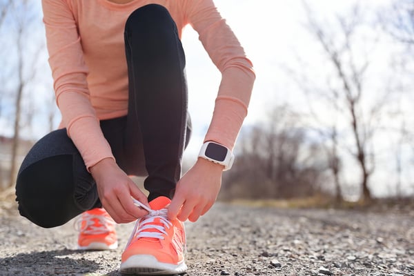 Runner with a white fitness tracker on one wrist kneels on a gravel path to tie orange running shoes