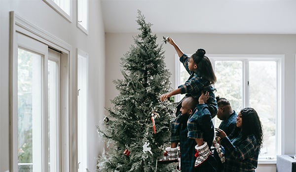 Girl on a man's shoulders placing a star atop a Christmas tree as others in matching plaid pajamas watch