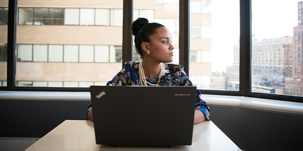 Woman with a laptop sits by a large office window overlooking city buildings