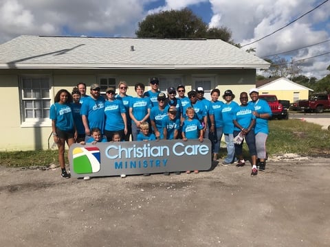 Adult and child volunteers in matching blue T-shirts pose behind a Christian Care Ministry sign on the Day of Sharing