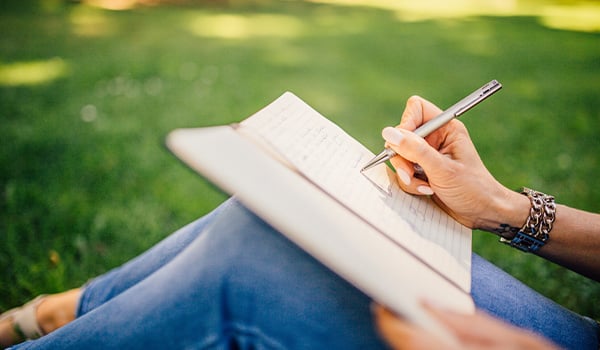 Person sitting on the grass writing in a notebook with a pen