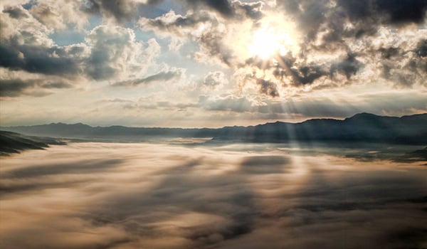 Sun breaking through clouds, casting rays over a fog-filled valley ringed by mountains