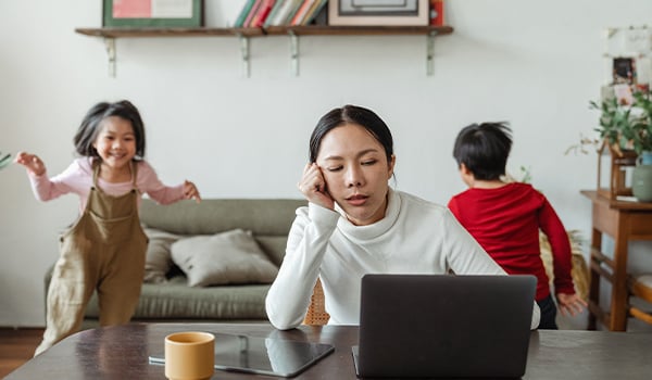 Tired woman resting her head on her hand at a laptop while two young children play behind her