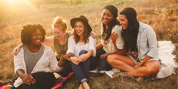 Five young women laughing with their arms around each other on a blanket in a sunlit field