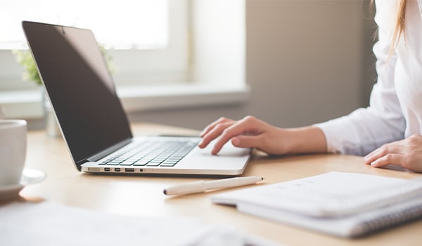 Person typing on a laptop at a sunlit desk with a notebook, pen and coffee cup
