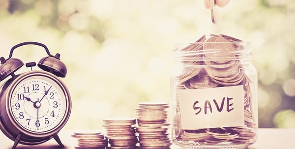 Hand dropping a coin into a glass jar labeled Save next to stacks of coins and an alarm clock
