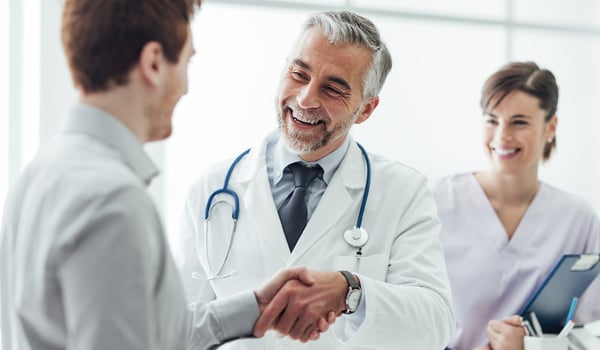 Smiling doctor in a white coat shakes hands with a patient as a nurse with a clipboard looks on