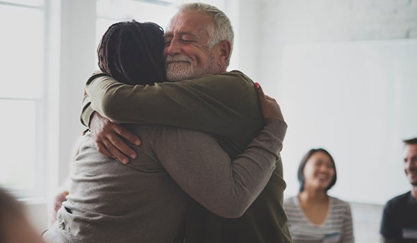 Smiling older man sharing a warm hug with another person as others look on in a bright room