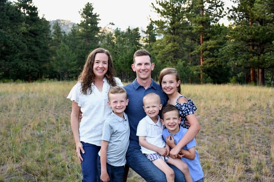 Smiling family with four children posing together in a grassy meadow in front of pine trees