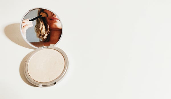 Open makeup compact with its mirror reflecting a young woman applying makeup with a brush