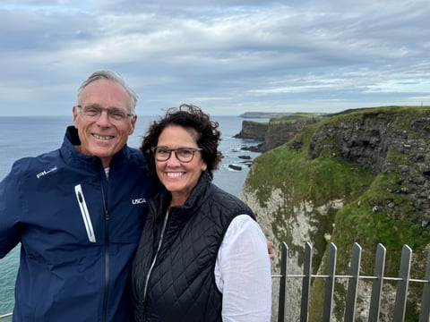 Smiling couple in glasses poses at a railing above green sea cliffs and the ocean