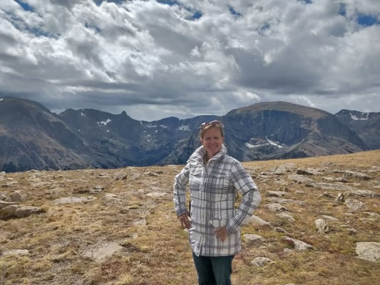Smiling woman in a plaid jacket on a rocky mountain plateau, with rugged peaks and clouds behind her