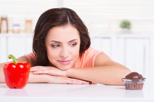 Woman resting her chin on her arms, eyeing a chocolate cupcake, with a red bell pepper on the other side