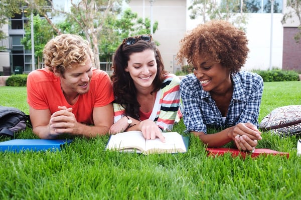 Three young adults lying on a campus lawn, smiling as they study an open book together
