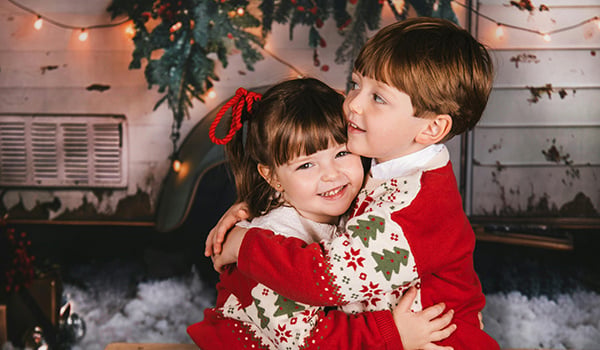 Two young children in matching Christmas sweaters hugging in front of string lights
