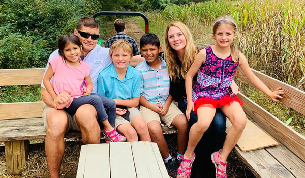 Two adults and four children smile on a wooden hayride wagon beside a cornfield