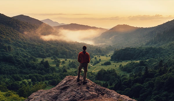 Man standing on a rocky ledge overlooking a misty forested valley and mountains in golden light