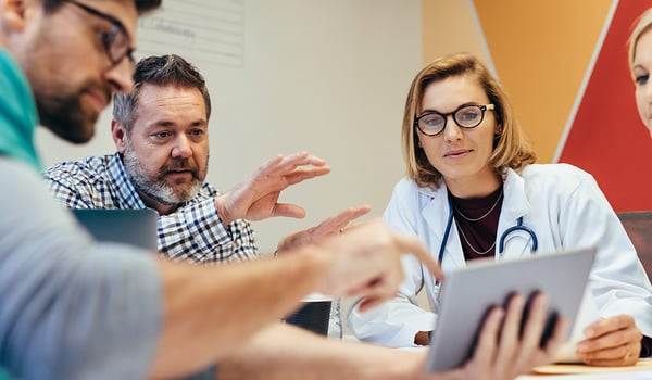 Doctor in a white coat and colleagues talk over a tablet at a meeting table