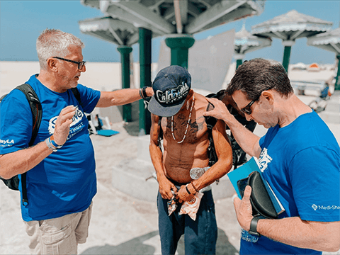 Two volunteers in matching blue T-shirts pray with a man wearing a California cap at a sunny beachfront