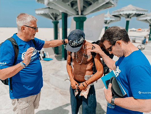 Two volunteers in matching blue T-shirts pray with a man wearing a California cap at a sunny beachfront