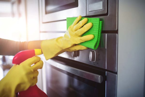 Gloved hands wiping a stainless steel oven with a green cloth while holding a spray bottle