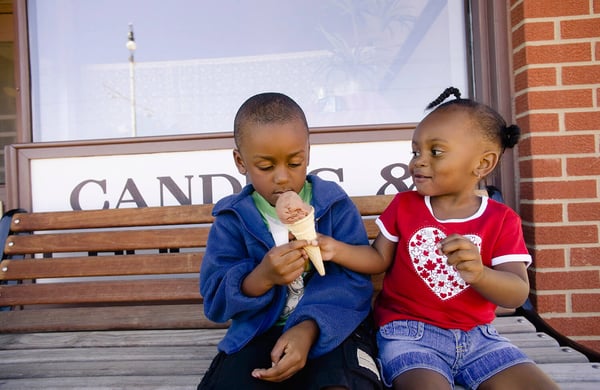 Two young children sharing an ice cream cone on a bench outside a candy shop
